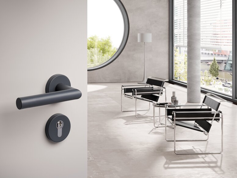 Dark grey lever handle with a round rose and matching keyhole escutcheon on a light grey door, with black armchairs and large windows in the bright room beyond