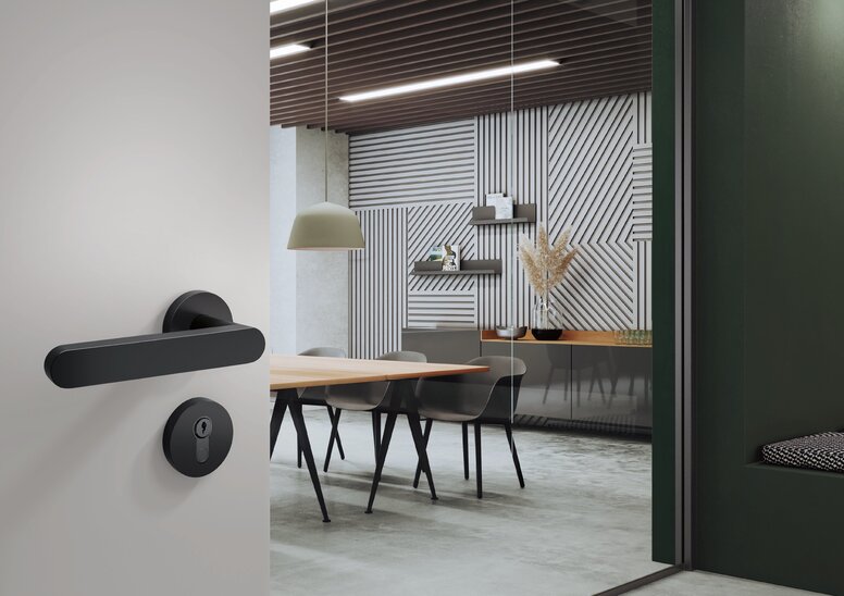 A black door handle with a round rose and a matching lock cylinder fitted to a light grey door; behind it is a meeting room