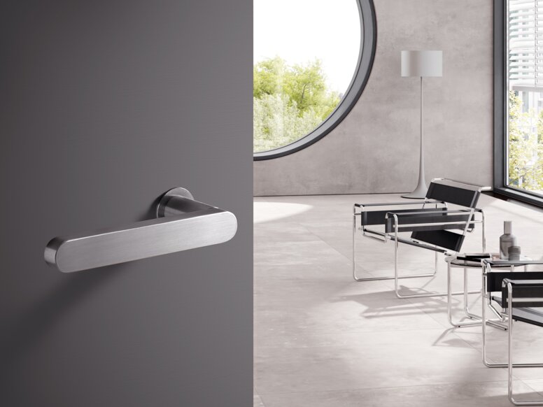 HEWI Range 270 lever handles A stainless steel door handle on a grey door; behind it, a bright room with chairs
