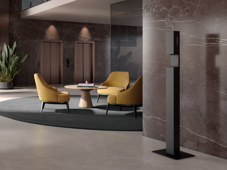 HEWI System 900 Disinfectant Dispenser Stand A free-standing black powder-coated hand sanitiser stand in a reception area in front of a brown marble wall