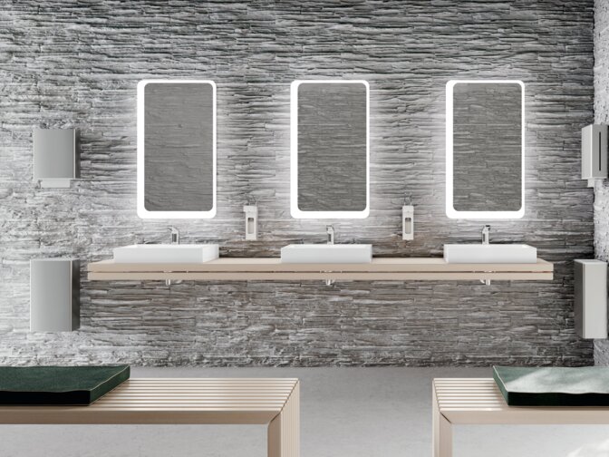Three rectangular illuminated mirrors above white countertop washbasins, each fitted with two soap dispensers and waste bins, in the public WC
